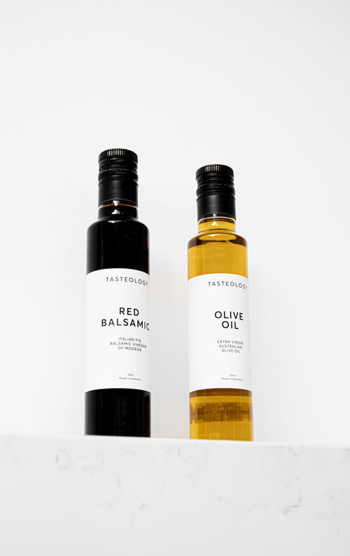 Two Tasteology bottles, Red Balsamic and Olive Oil, standing side-by-side on a white marble surface against a white background.