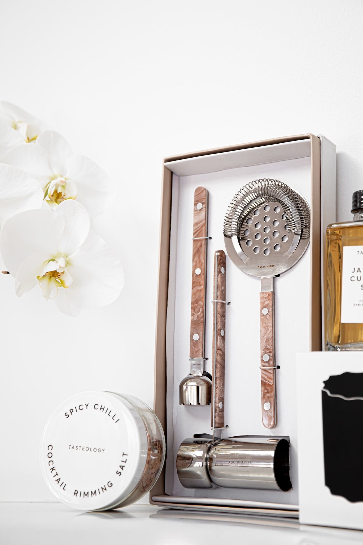A bar tool set in an open presentation box, including a strainer, muddler, spoon, and chiller, with woodgrain handles and silver metal heads. The box is styled on a white surface with a white orchid flower, a round tin labeled 'Spicy Chilli Cocktail Rimming Salt', and part of a bottle visible.