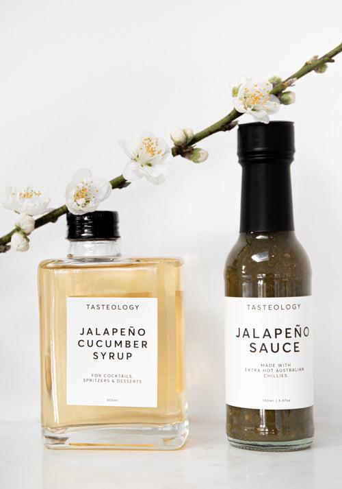 Two Tasteology condiment bottles on a white surface: a square glass bottle of jalapeño cucumber syrup with pale golden liquid and a slim glass bottle of jalapeño sauce with dark green sauce inside, both with black caps and white labels, with white blossoms across the top.