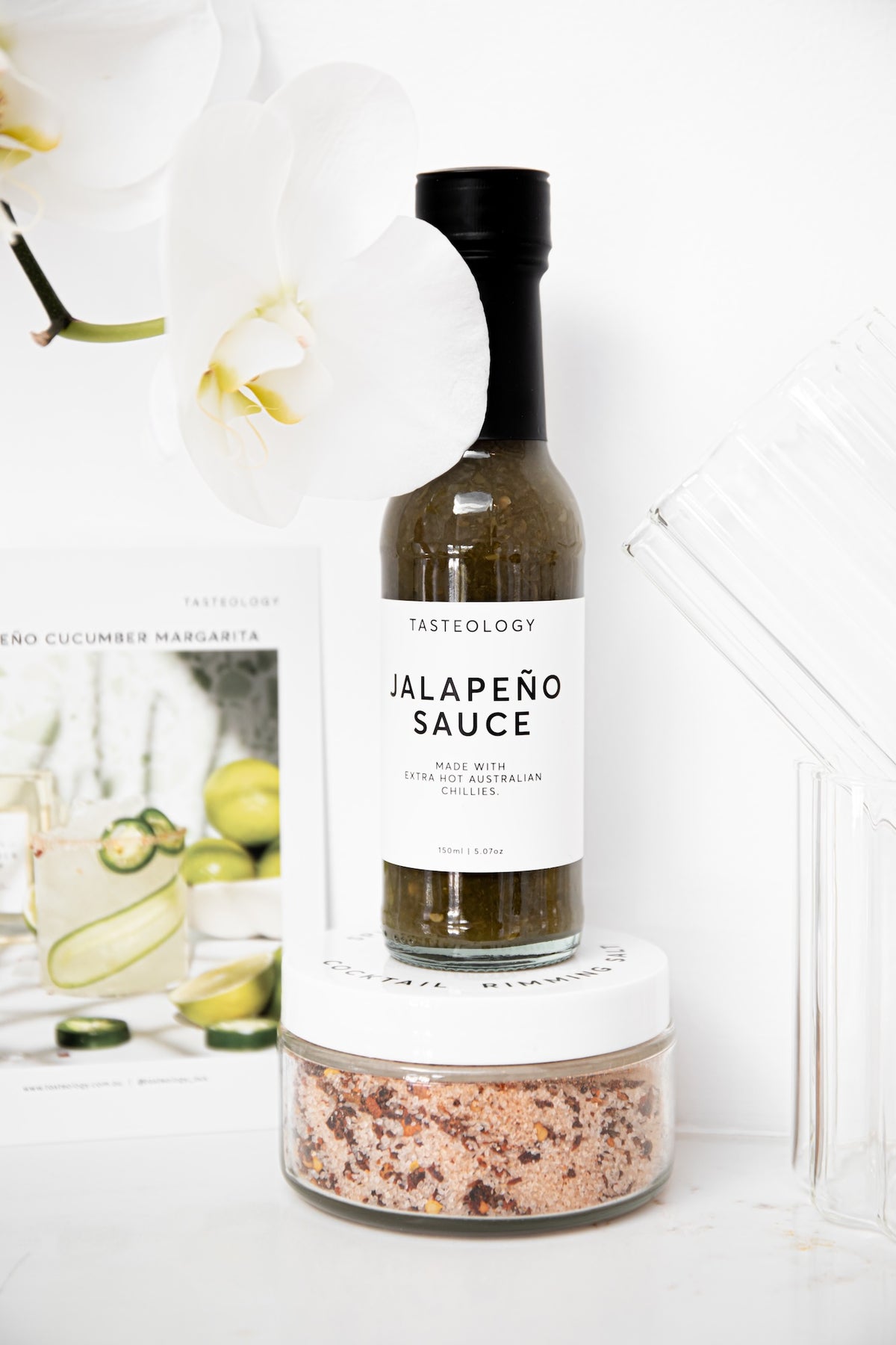 A bottle of Tasteology Jalapeño Sauce stands on top of a small clear jar filled with coarse salt, styled on a white surface with white orchid flowers and a magazine in the background.