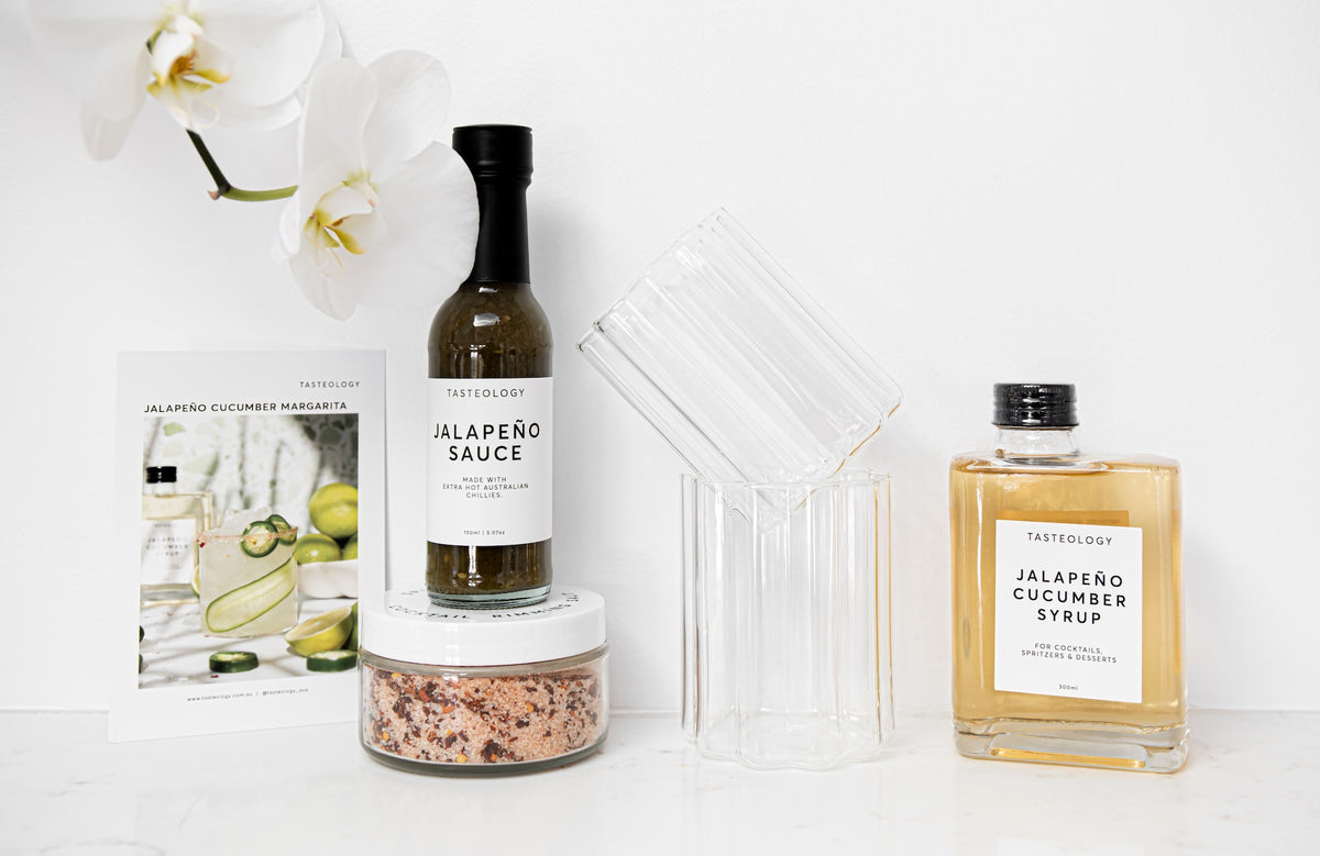 A styled product arrangement on a white surface featuring a bottle of TasteGroup Jalapeño Sauce, a bottle of TasteGroup Jalapeño Cucumber Syrup, a clear ribbed glass, a clear ribbed glass lid, a small jar of coarse seasoning, and a printed card, all photographed against a clean white background with white orchid flowers at the top.