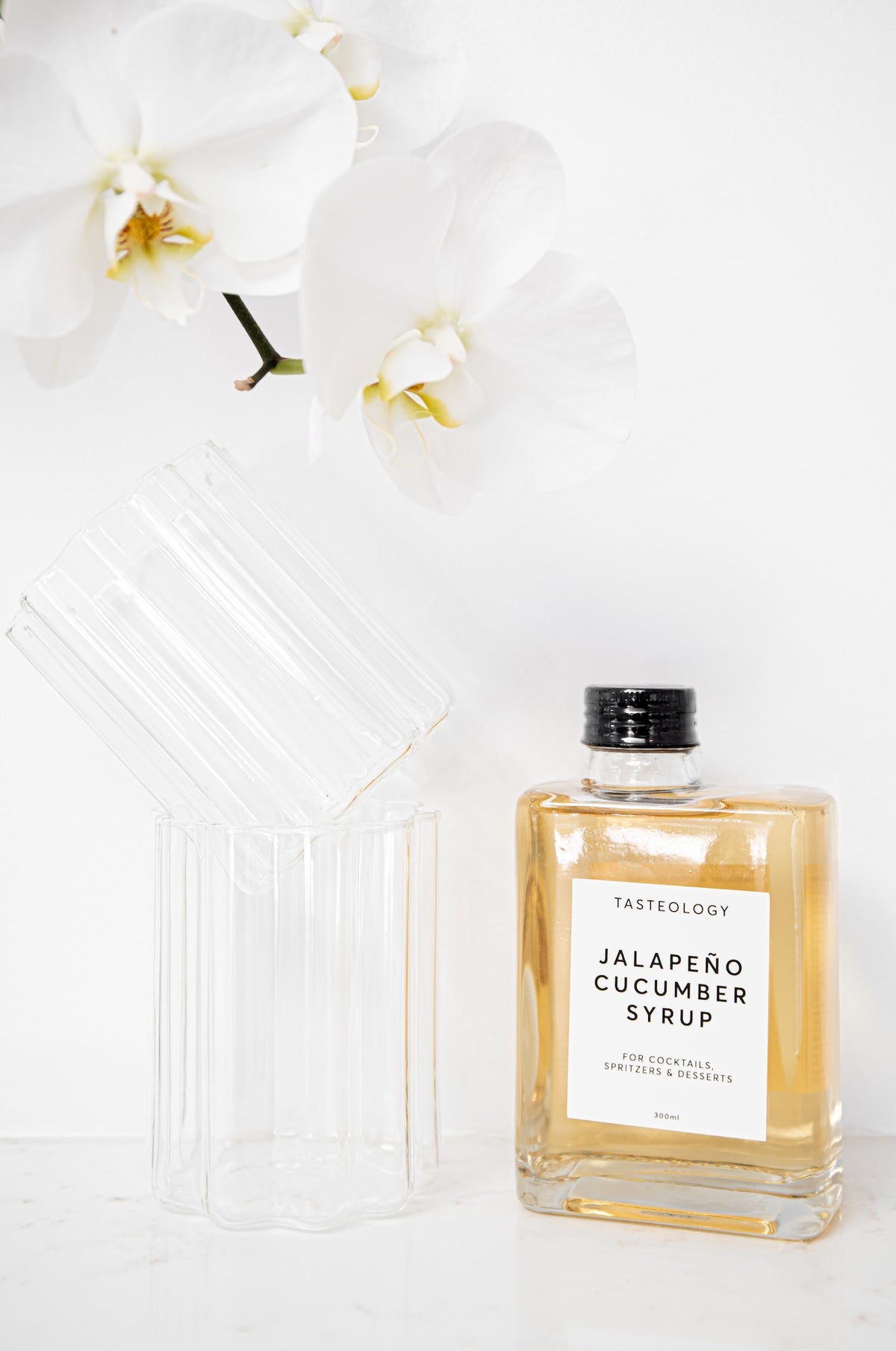 A bottle of Tasteology Jalapeño Cucumber Syrup with a clear ribbed glass container and a white label, shown next to a matching ribbed glass lid on a white surface with white orchids in the background.
