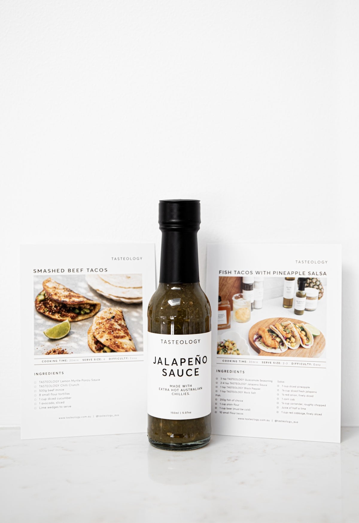 A bottle of Tasteology Jalapeño Sauce stands centered on a white surface, flanked by two printed recipe cards showing prepared tacos and salsa ingredients.