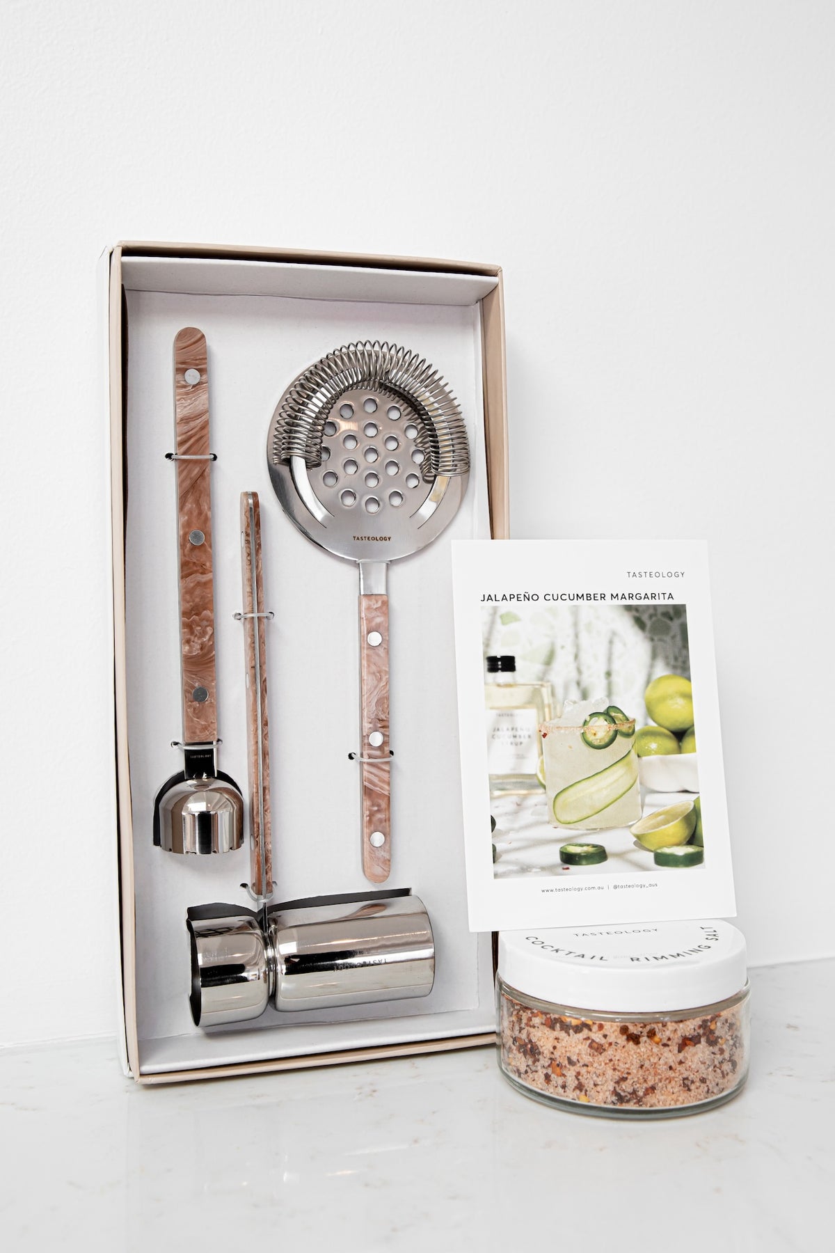 A cocktail bar gift set displayed in an open box, featuring a strainer, muddler, ice paddle, and bitters holder with wood-and-stone handles and polished metal ends. A small printed card and a jar of salt are placed beside the box on a clean white surface.