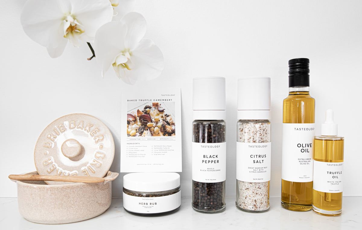 A styled product lineup of gourmet food items on a white surface against a white background, featuring a ceramic truffle dish, a recipe card, a jar of sea salt, a bottle of black pepper, a bottle of citrus salt, a bottle of olive oil, and a bottle of truffle oil.