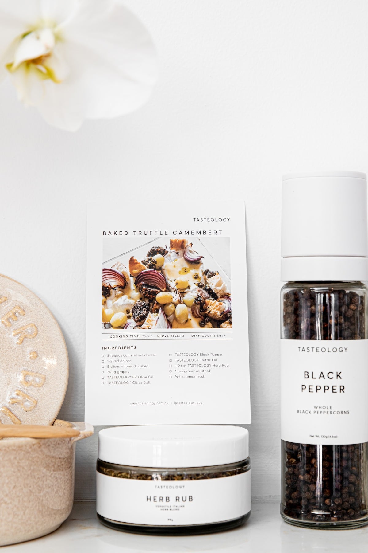 A styled product shot featuring Tasteology Baked Truffle Camembert ingredients: a jar of Herb Rub, a bottle of Black Pepper, and a recipe card, arranged on a white surface next to a ceramic dish with a white orchid flower in the background.