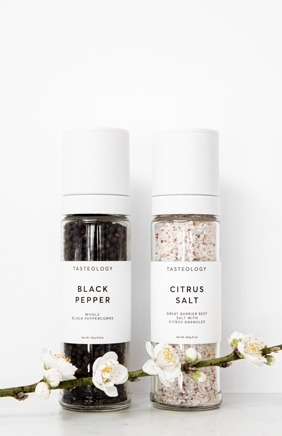 Two Tasteology spice grinders, one labeled Black Pepper and the other Citrus Salt, standing side by side on a white surface with white blossoms in the foreground against a white background.