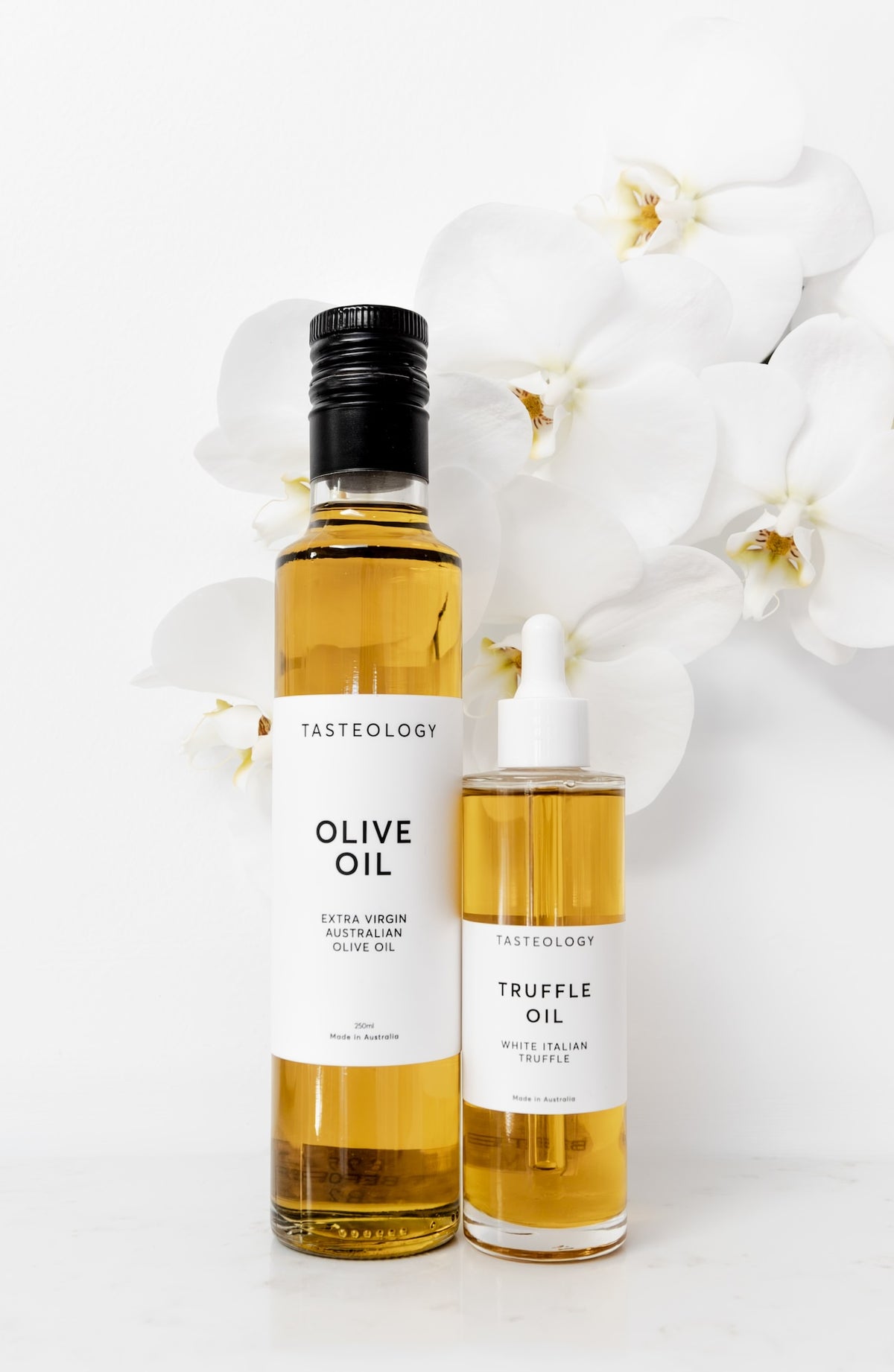 Two Tasteology oil bottles on a white surface with white orchid flowers behind them: a tall bottle of olive oil with a black cap and a shorter dropper bottle of truffle oil with a white dropper top.