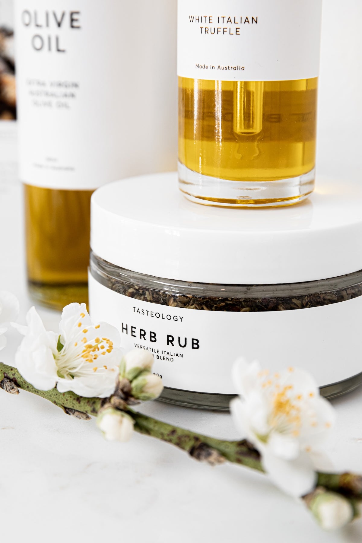 A styled product shot featuring a jar of Tasteology Herb Rub truffle rub in the foreground, with a bottle of White Italian Truffle oil behind it and white blossoms in the foreground, all on a light marble surface.
