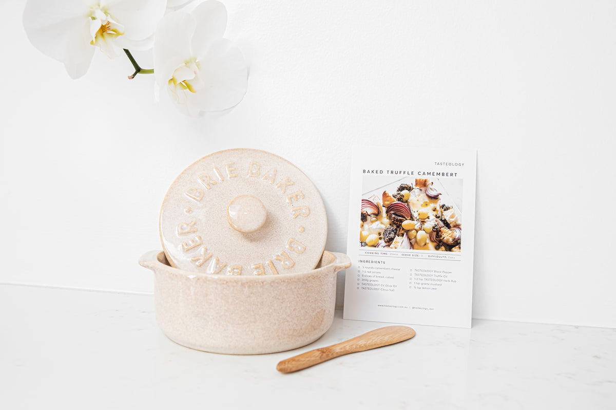 A speckled beige ceramic lidded baking dish with embossed text, shown beside a printed recipe card and a small wooden spatula on a white surface against a white wall.