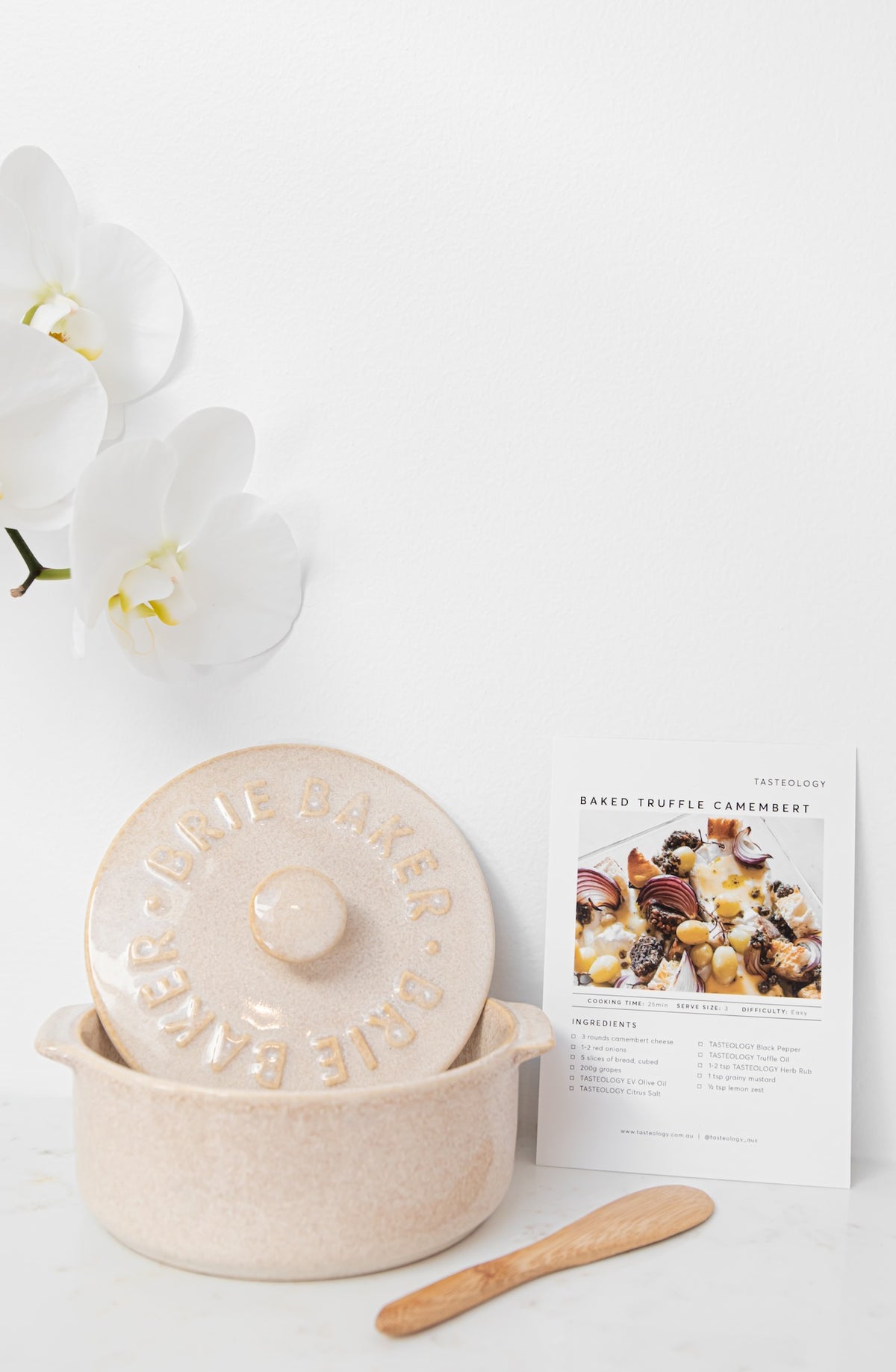 A ceramic truffle keeper pot with its lid on, shown on a white marble surface with white orchids and a recipe card. The pot and lid are a speckled cream-beige color, and a small wooden spatula rests in front.