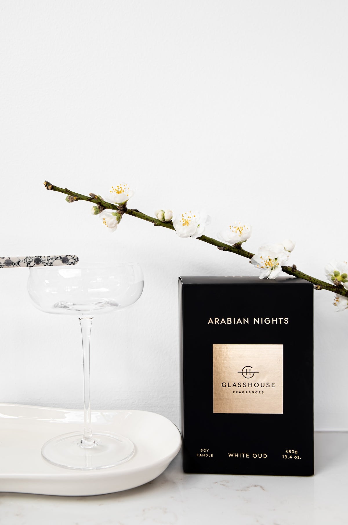 A black product box labeled 'ARABIAN NIGHTS' and 'GLASSHOUSE FRESHENING' stands on a white surface beside a clear stemmed glass and a white plate, with a flowering branch across the top against a white background.