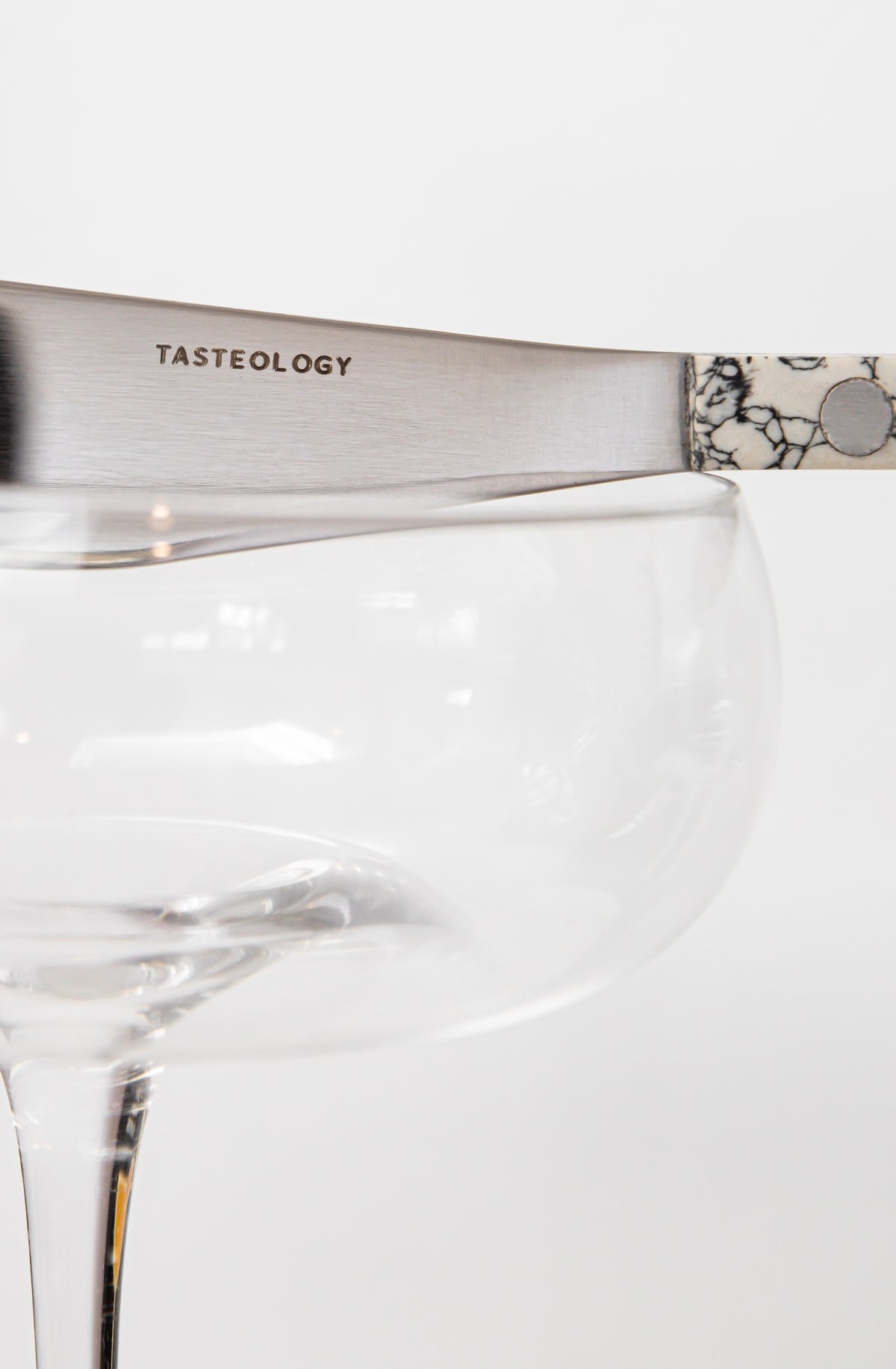 A close-up of a silver metal knife with the word 'TASTEOLOGY' engraved on the blade, resting on the rim of a clear glass coupe glass against a white background.