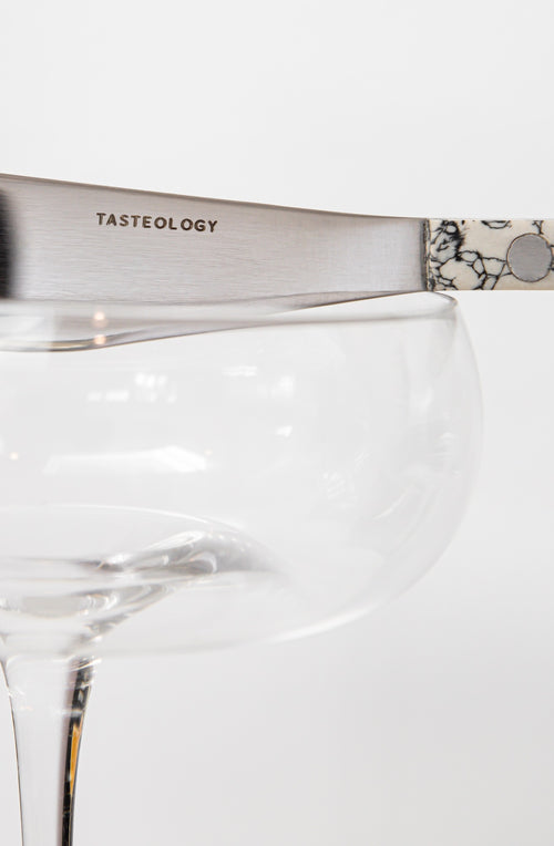 A close-up of a silver metal knife with the word 'TASTEOLOGY' engraved on the blade, resting on the rim of a clear glass coupe glass against a white background.