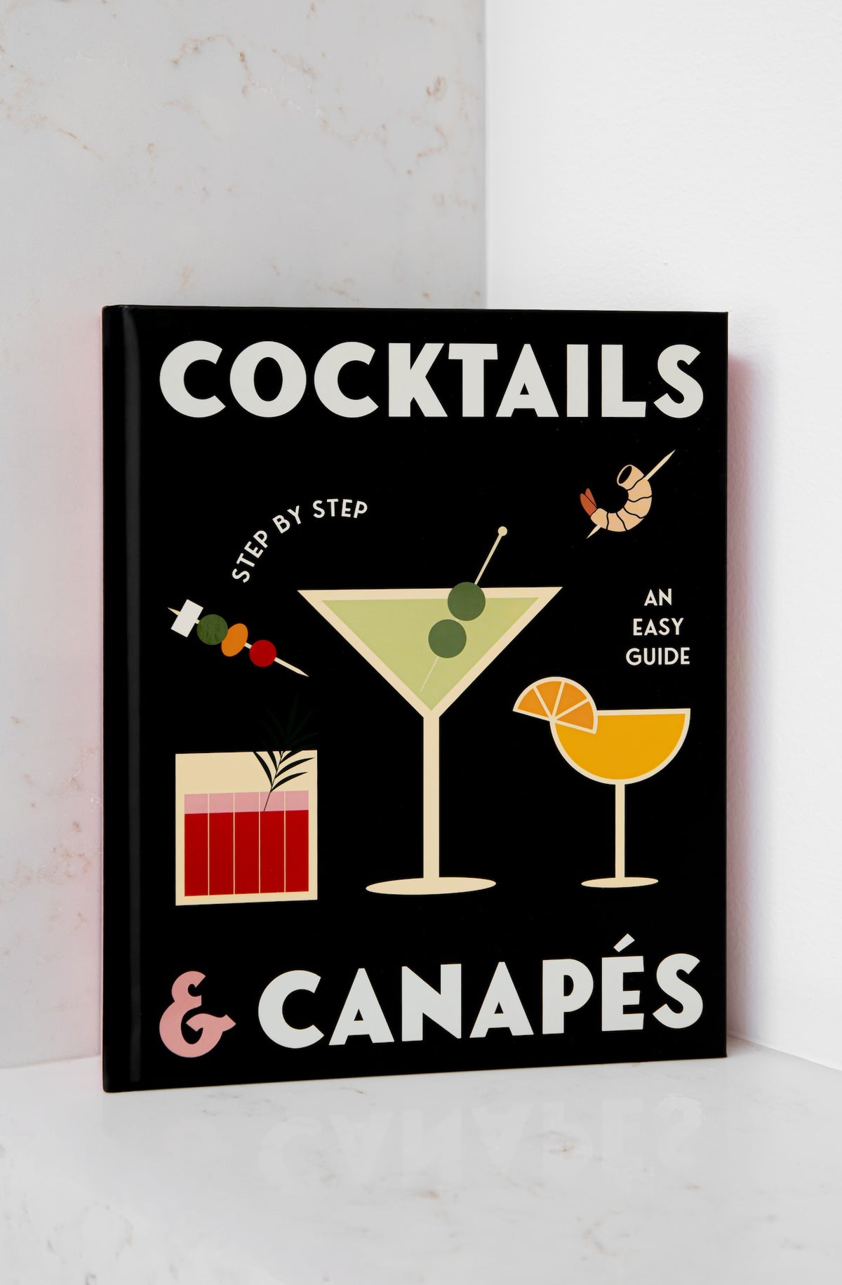 A hardcover book titled 'COCKTAILS & CANAPÉS: AN EASY GUIDE' with a black cover, featuring colorful illustrations of cocktails and canapes on a white marble surface.