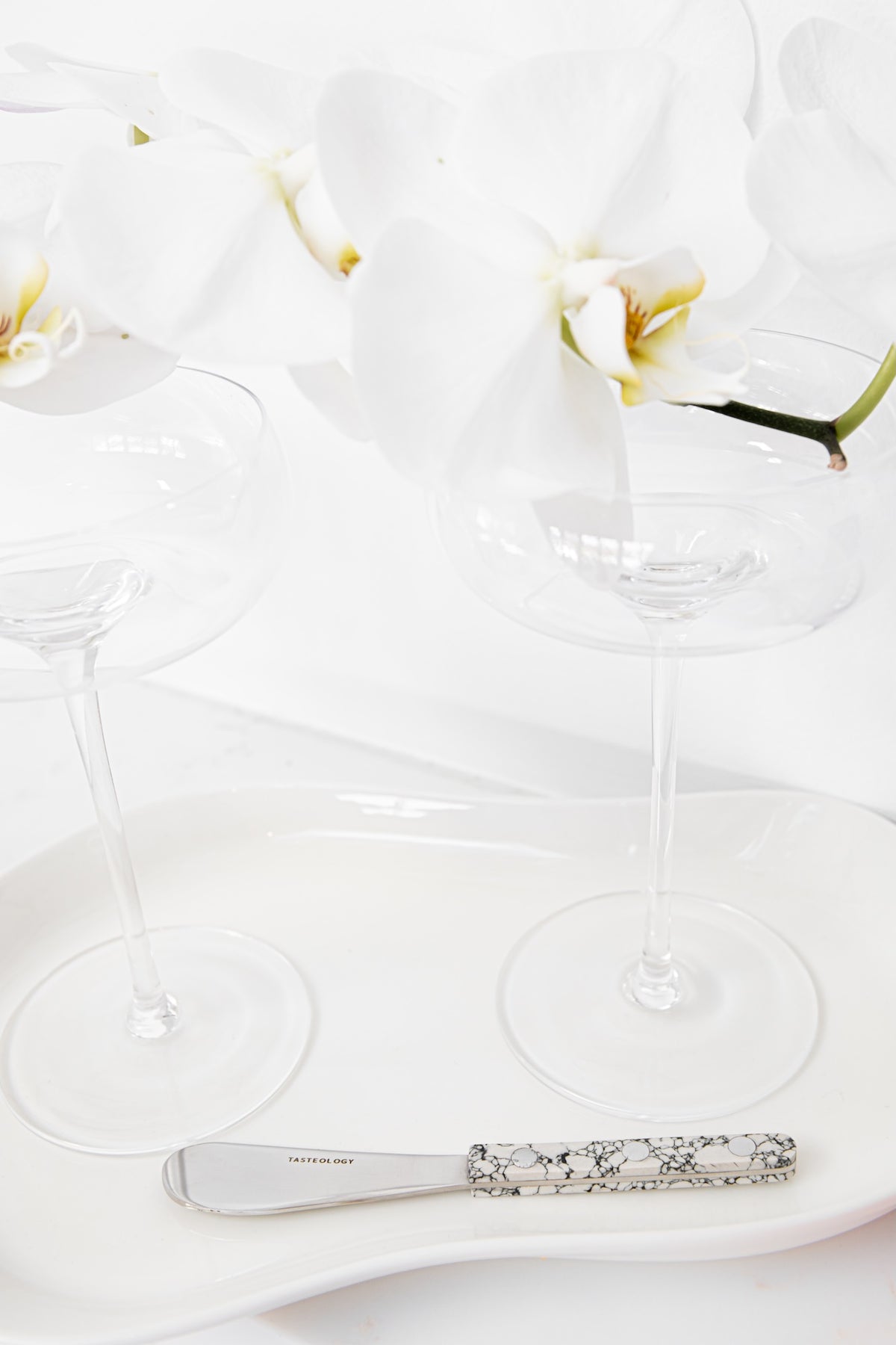 A close-up of a silver butter knife with an ornate floral handle resting on a white ceramic tray, with two clear wine glasses and white orchids in the background.