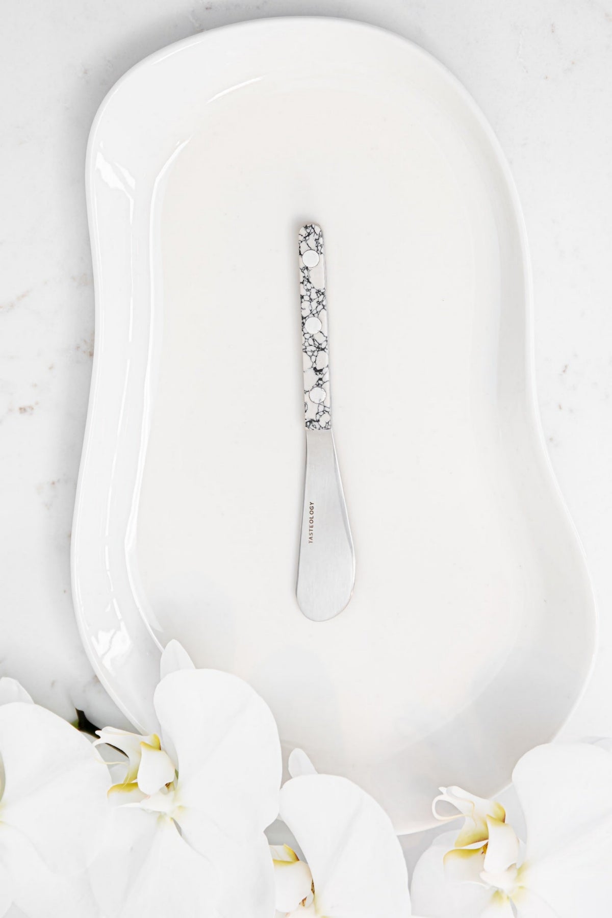 A white ceramic serving platter with a decorative silver butter knife featuring a black floral pattern handle, styled on a white marble surface with white orchids.