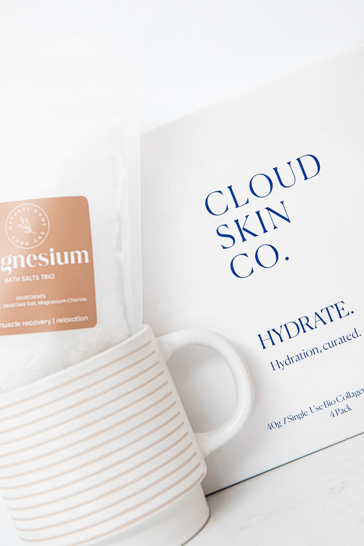 Cloud Skin Co. Hydrate bath salts in a white bag with a blue logo, shown next to a white ceramic mug with thin gold stripes on a light marble surface.