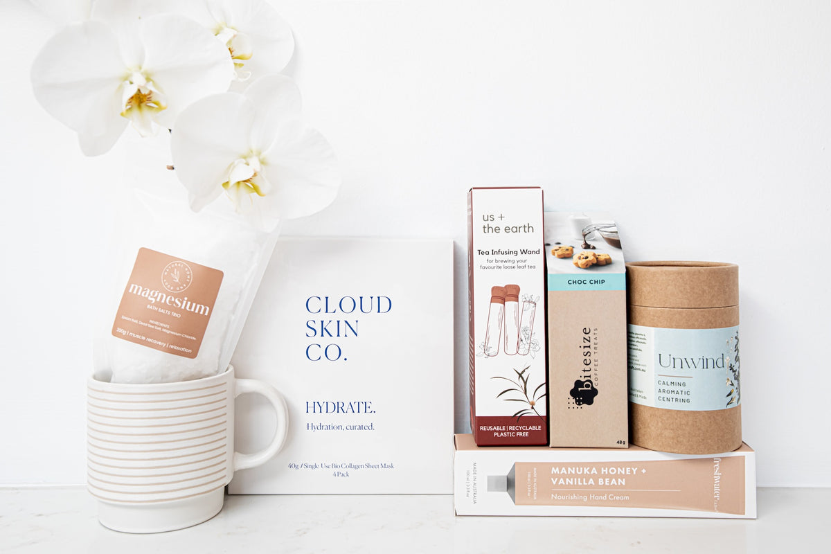 A styled flat-lay of multiple wellness and skincare products on a white surface against a white background, including Cloud Skin Co. Hydrate tea, a mug with a magnesium sachet, a tube of vanilla bean honey, and other boxed items.