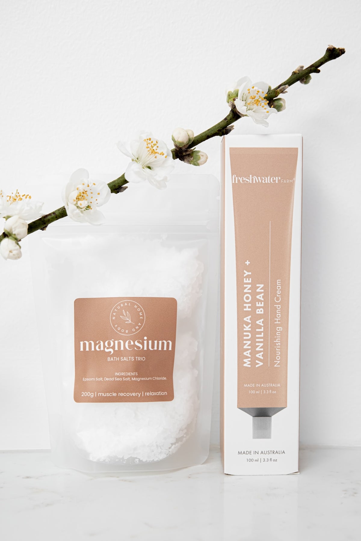 A skincare set featuring a clear stand-up pouch of magnesium bath salts and a matching tube of Freshwater Manuka Honey + Vanilla Bean moisturising hand cream, styled with white blossoms on a clean white background.