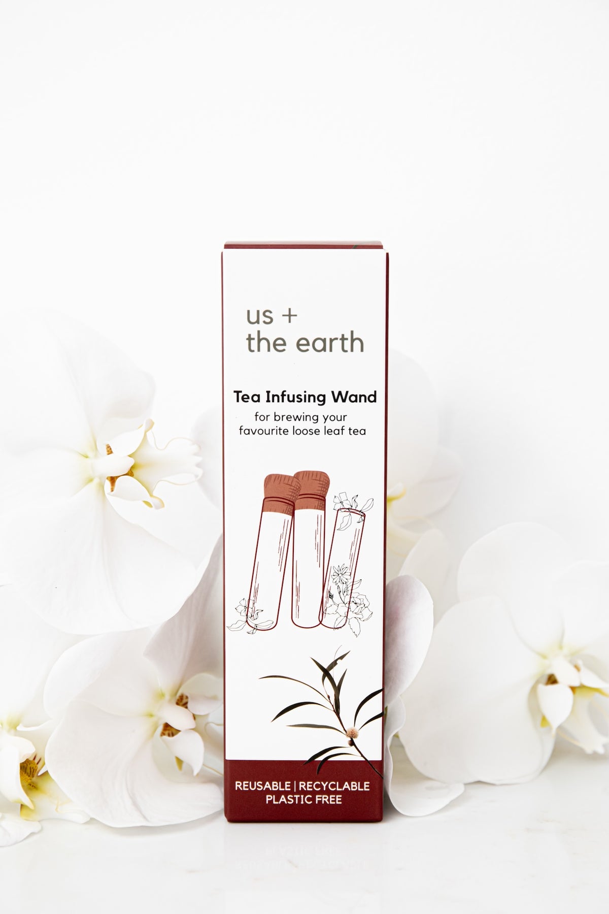 A boxed tea infusing wand by us + the earth, standing upright on a white surface with white orchid flowers around it. The box is white with dark red bands and printed illustrations of two glass infusing wands.