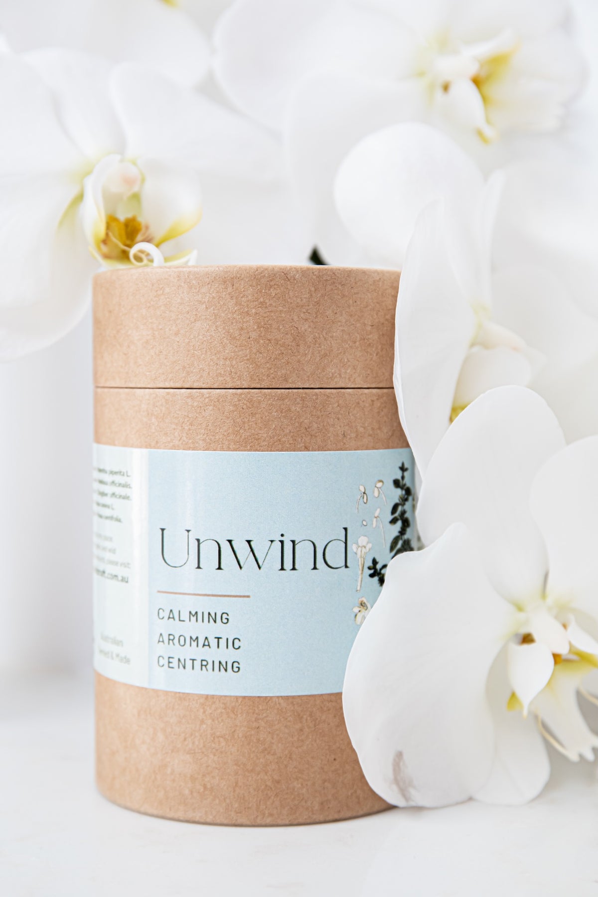 A kraft paper cylindrical container with a light blue label reading 'Unwind Calming Aromatic Centring,' styled with white orchid flowers on a soft white background.