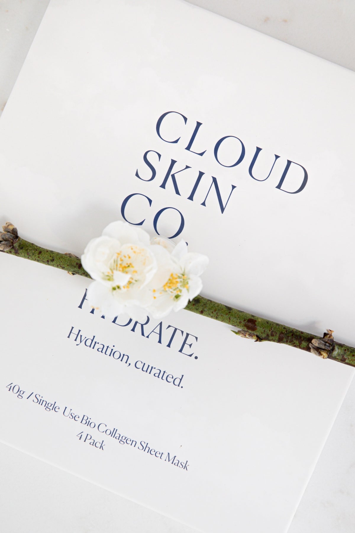 Cloud Skin Co. Hydrate Collagen Sheet Mask packaging, featuring white paper sleeves with navy blue text and a decorative white flower resting on top.