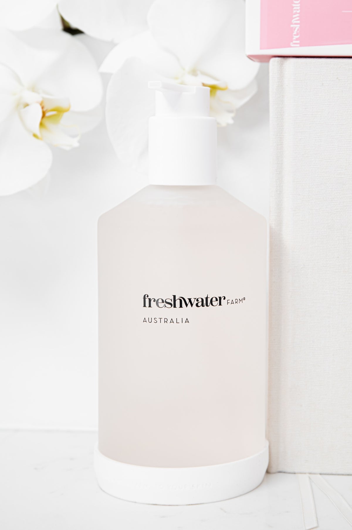 A frosted glass pump bottle of Freshwater Farm Australia liquid, shown with a white base and pump, styled with white orchids and a white textured box in the background.