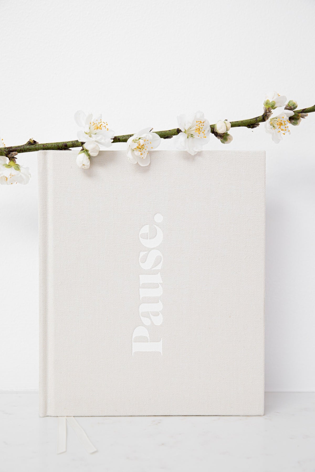 A square hardcover journal with a textured off-white linen cover, featuring the word 'Pause.' printed vertically in a matching light tone.