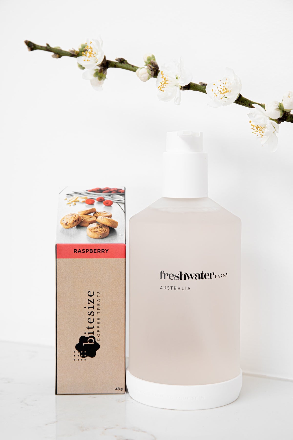 A frosted bottle of Freshwater by Bitesize Australia raspberry mouthwash next to its kraft paper packaging box, styled with a white background and a flowering branch.