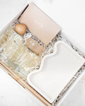 A luxury gift set in a white box, featuring a clear glass perfume bottle with a wooden cap, a small wooden diffuser, a beige card labeled 'sainte', and a white ceramic tray, arranged on white shredded paper.