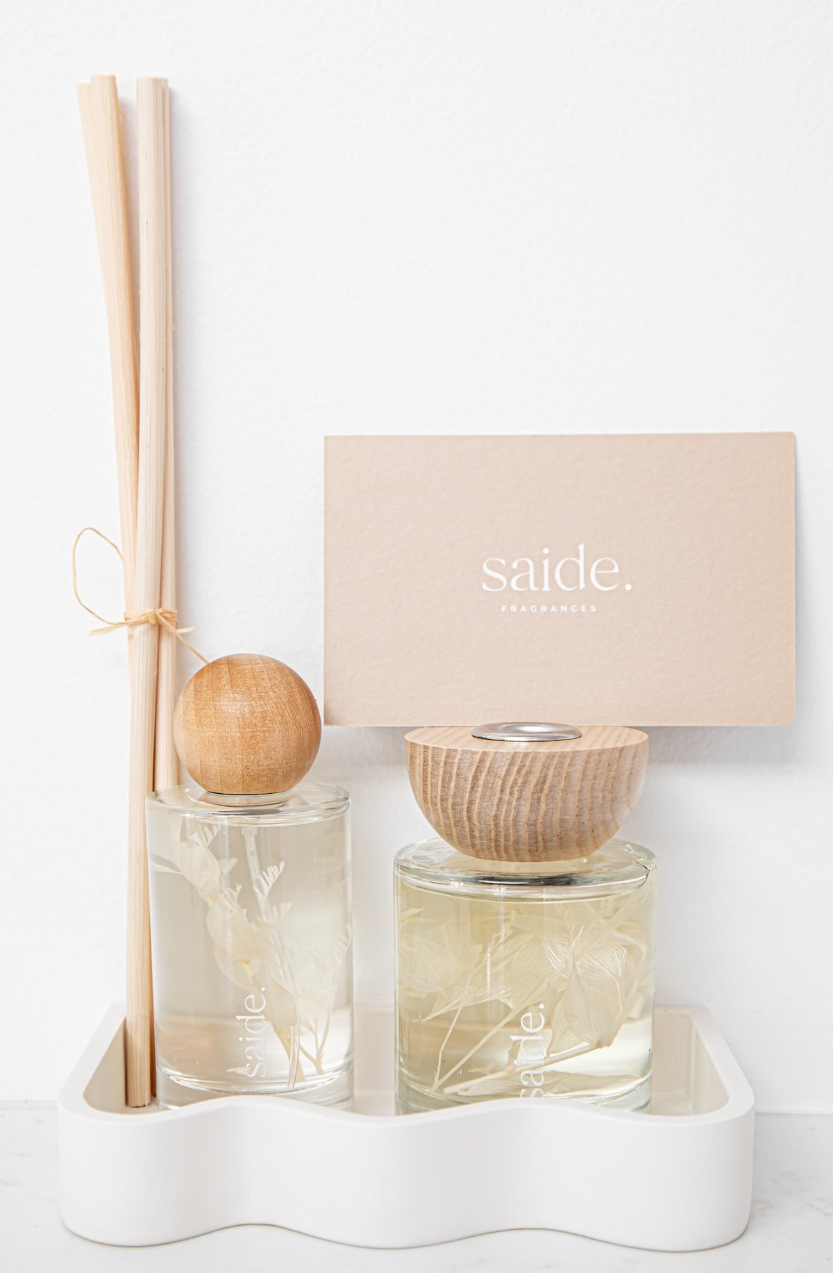 A reed diffuser set with two glass bottles, one with a wooden cap and one with a reed stick in a white holder, alongside a beige card printed with the brand name 'saide'.