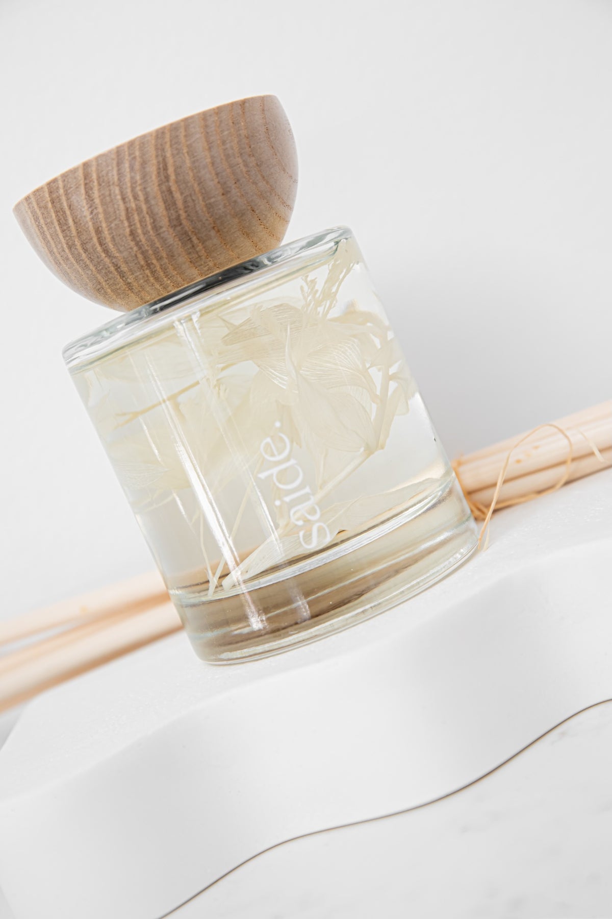 A clear glass reed diffuser bottle with a light wood bowl cap, filled with pale liquid and reeds, shown on a white surface with a soft neutral background.