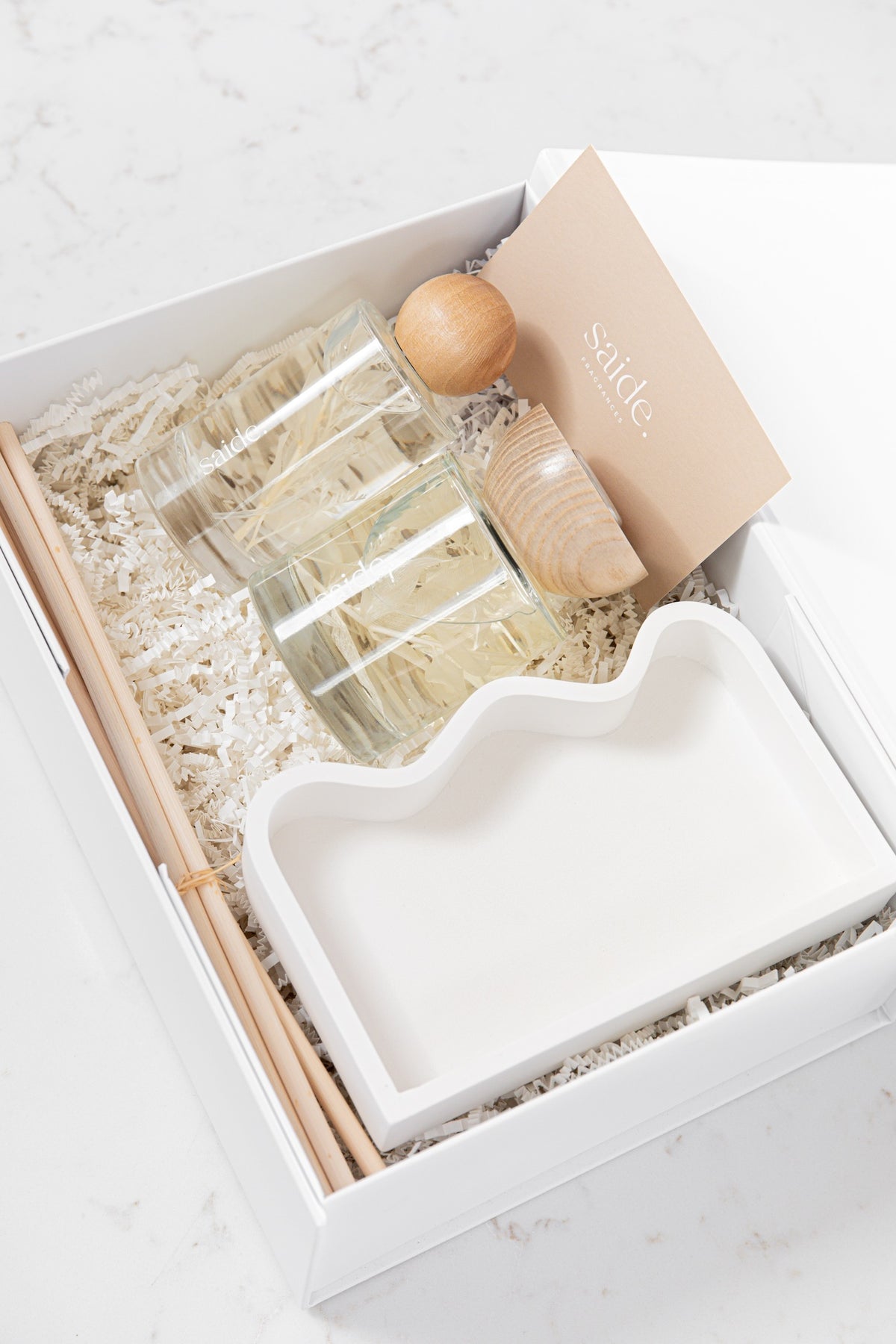 A white gift box containing a clear glass diffuser bottle, a white tray, wooden accessories, and a beige card with the brand name 'saide' on a light marble background.