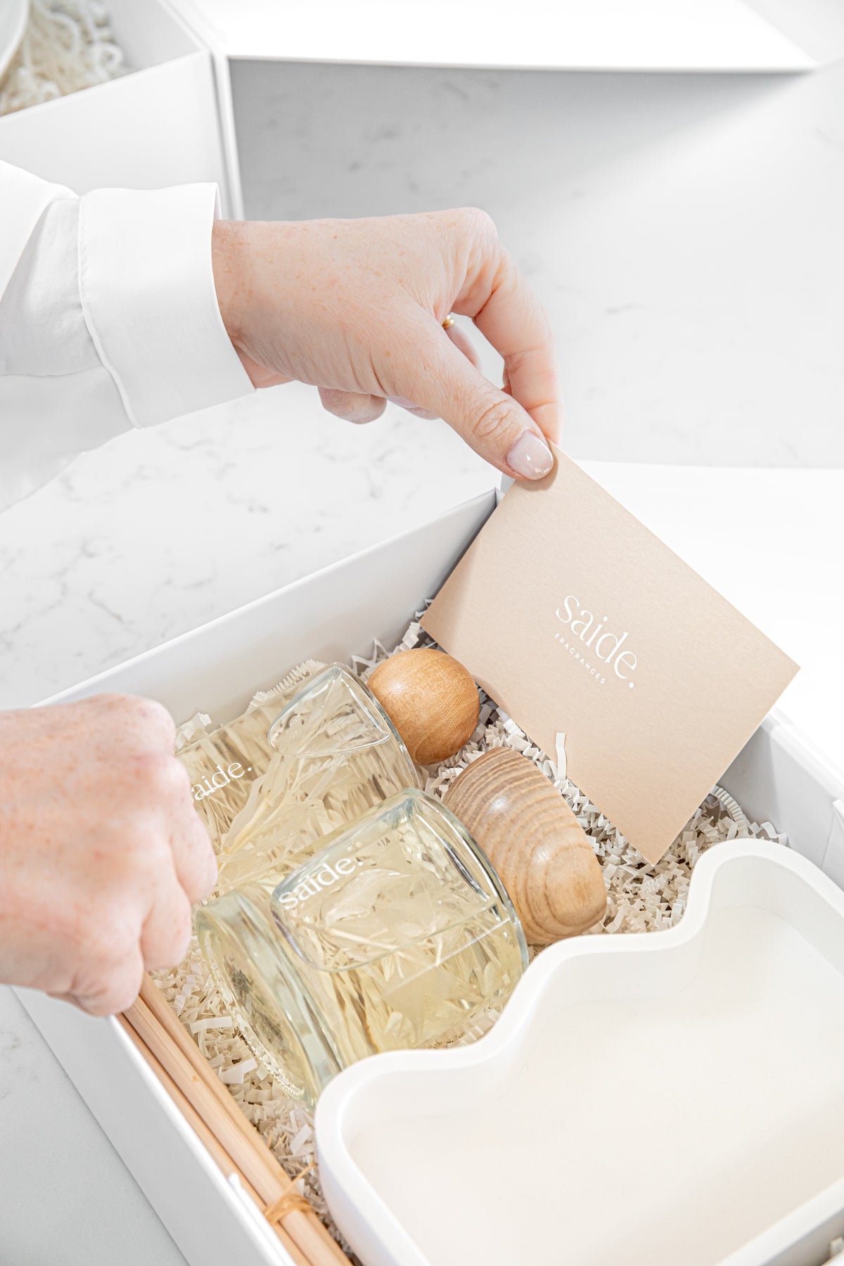 A close-up of hands opening a white gift box containing a glass reed diffuser bottle, wooden diffuser pieces, a small card labeled 'sainte', and a white cloud-shaped dish on a light marble surface.