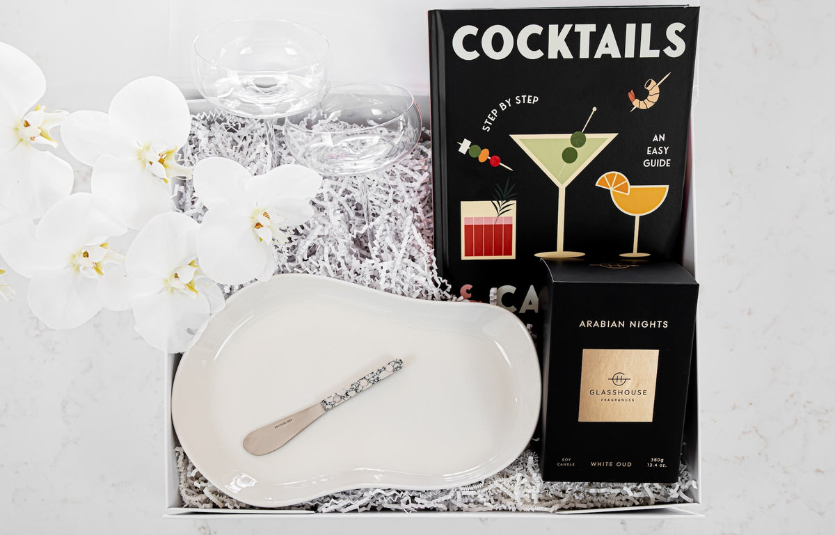 A cocktail-themed gift box set featuring a black 'COCKTAILS' book, a black 'ARABIAN NIGHTS' box, a white oval serving platter with a silver spoon, and clear glassware, arranged in white shredded paper with white orchids on a light marble surface.