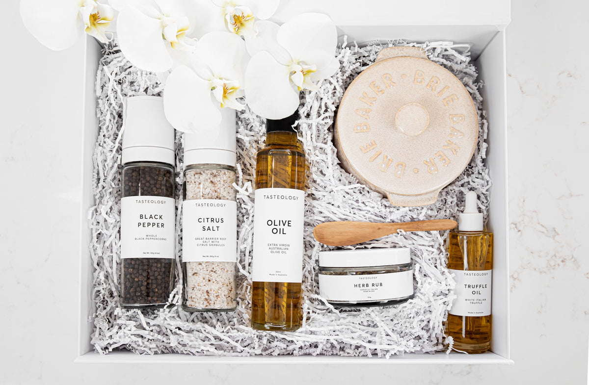 A curated gift box set from Tasteful Living, arranged in a white box with white paper shred. The set includes a bottle of black pepper salt, a bottle of citrus salt, a bottle of olive oil, a round Brie Baker