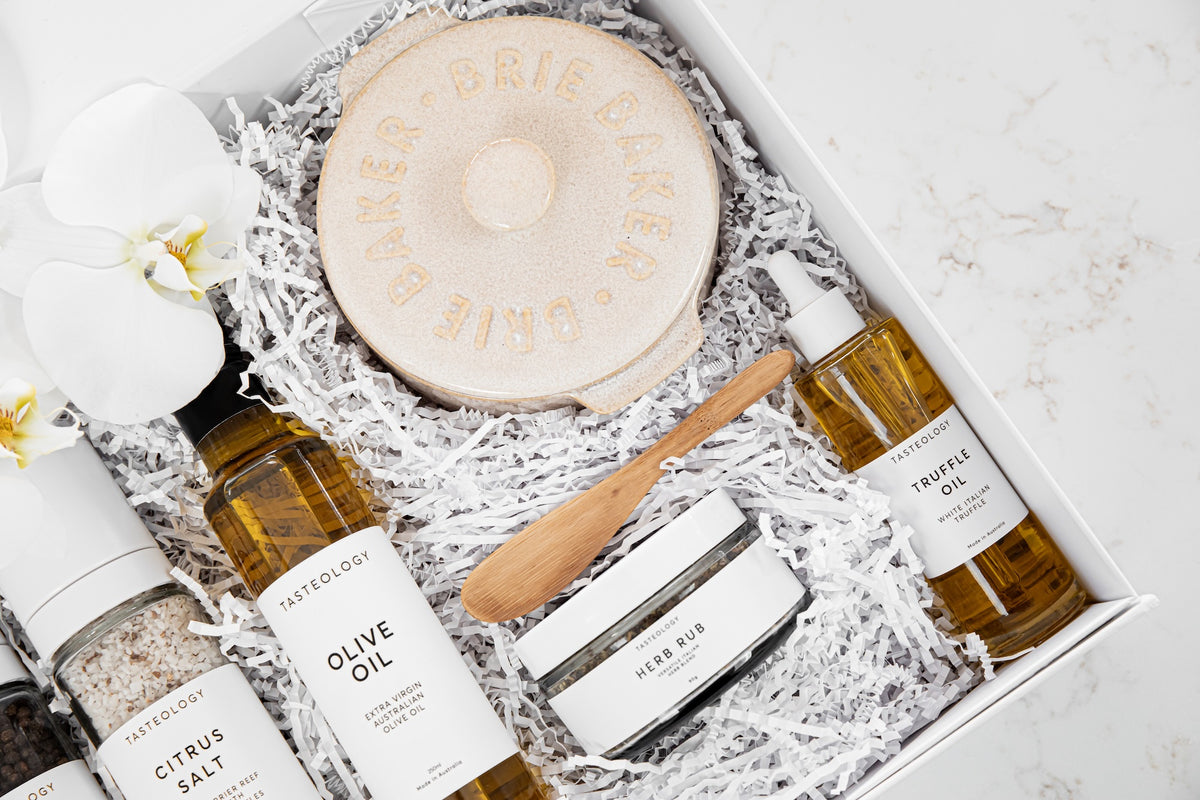 A curated gift box set from Attico Logic, featuring a round wooden board engraved with 'BRIE DAKER', olive oil, truffle oil, citrus salt, a cream-colored body butter jar, and a small wooden spatula, all nestled in white shredded paper inside a white box with a white orchid flower.