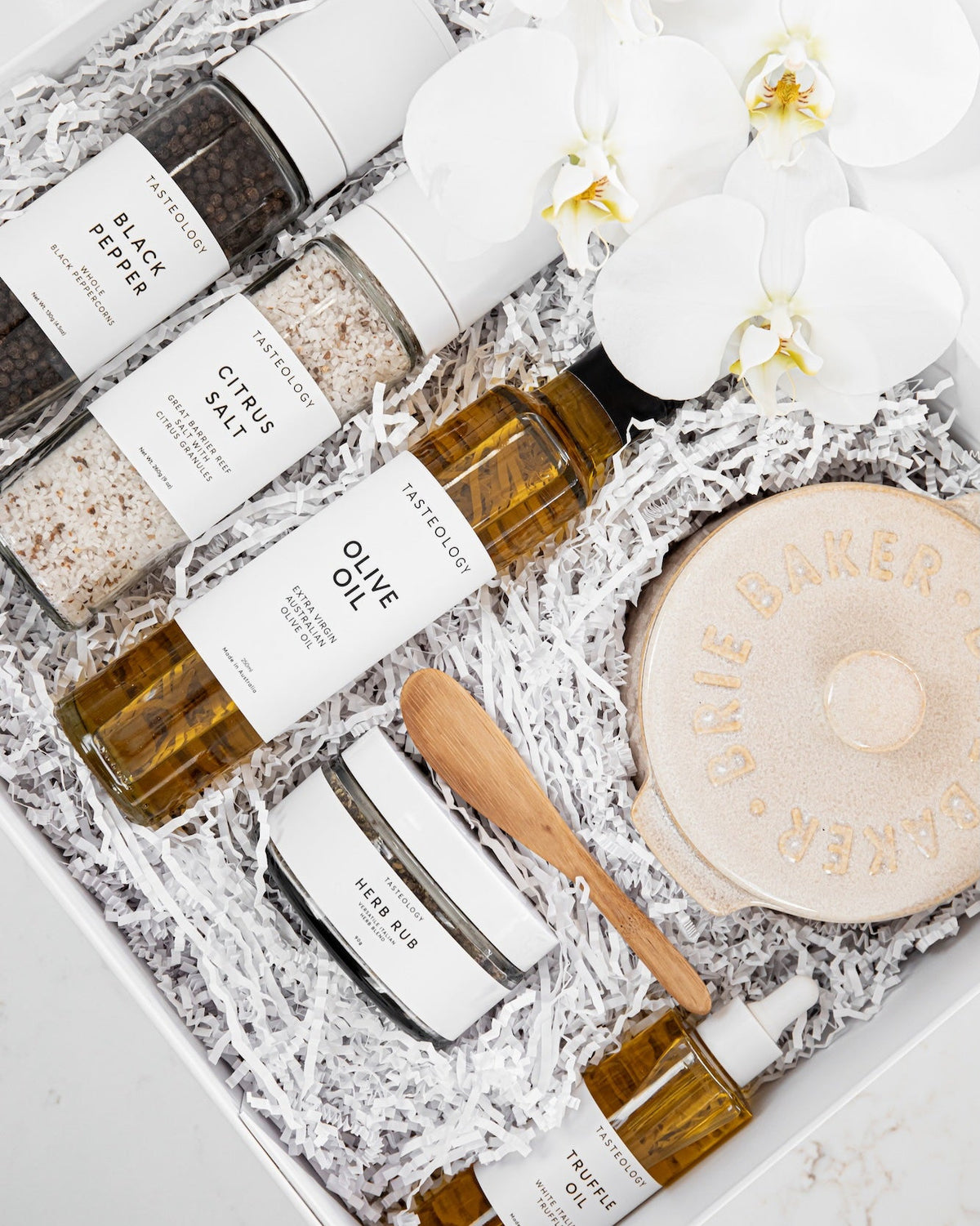 A curated gift box of gourmet food and beverage products arranged in white crinkle paper. The set includes bottles of olive oil, peppercorns, sea salt, citrus salt, honey, and a round wooden spoon, alongside a beige ceramic container embossed with 'BRIE BAKER'.
