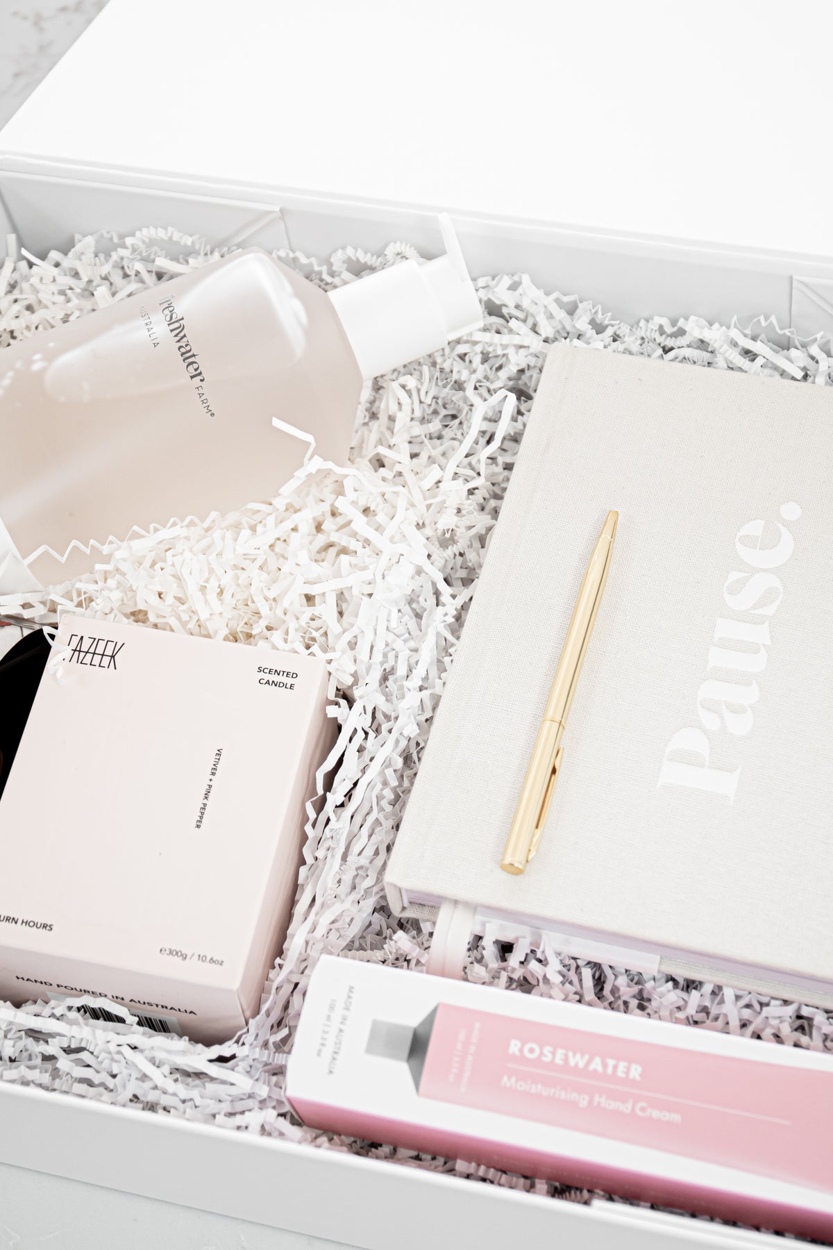 A luxury gift set arranged in a white box filled with white shredded paper. The set includes a frosted bottle, a pink box, a pink tube of moisturizing hand cream, a beige book or notebook with the word 'Pause.