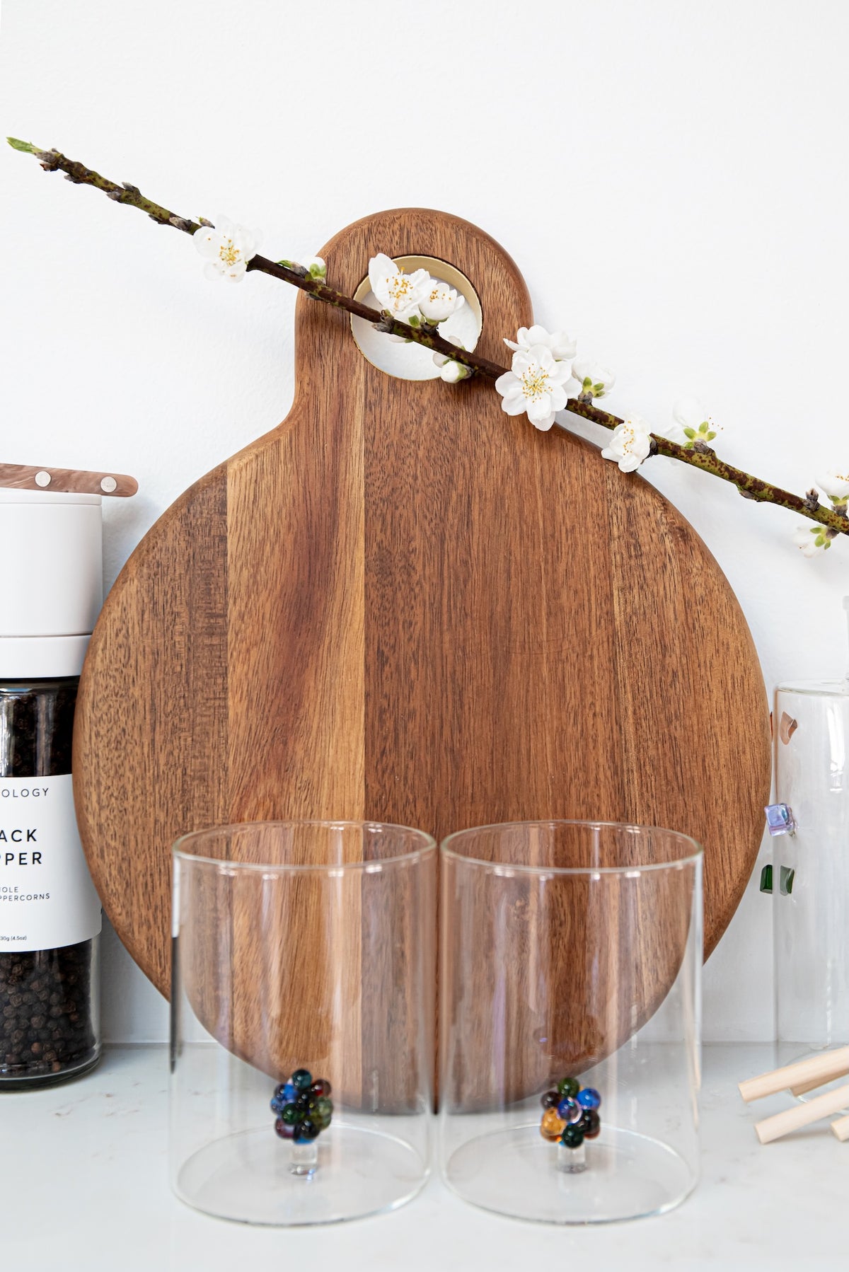 A round wooden cutting board with a handle and a floral blossom draped across the top, styled on a white surface with two clear glass tumblers in front and kitchen items partially visible at the sides.