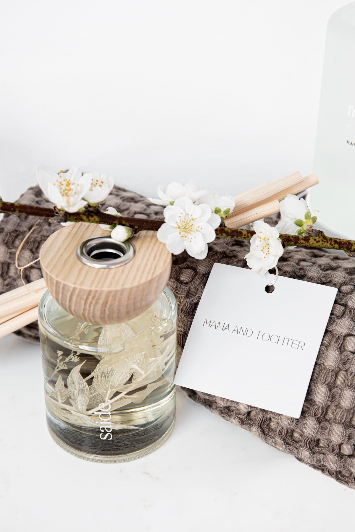 A glass reed diffuser bottle with a light wood cap, filled with clear liquid and dried botanical elements, styled on a textured gray blanket with white blossoms and a hanging tag reading 'MAMA AND TOCHTER'.