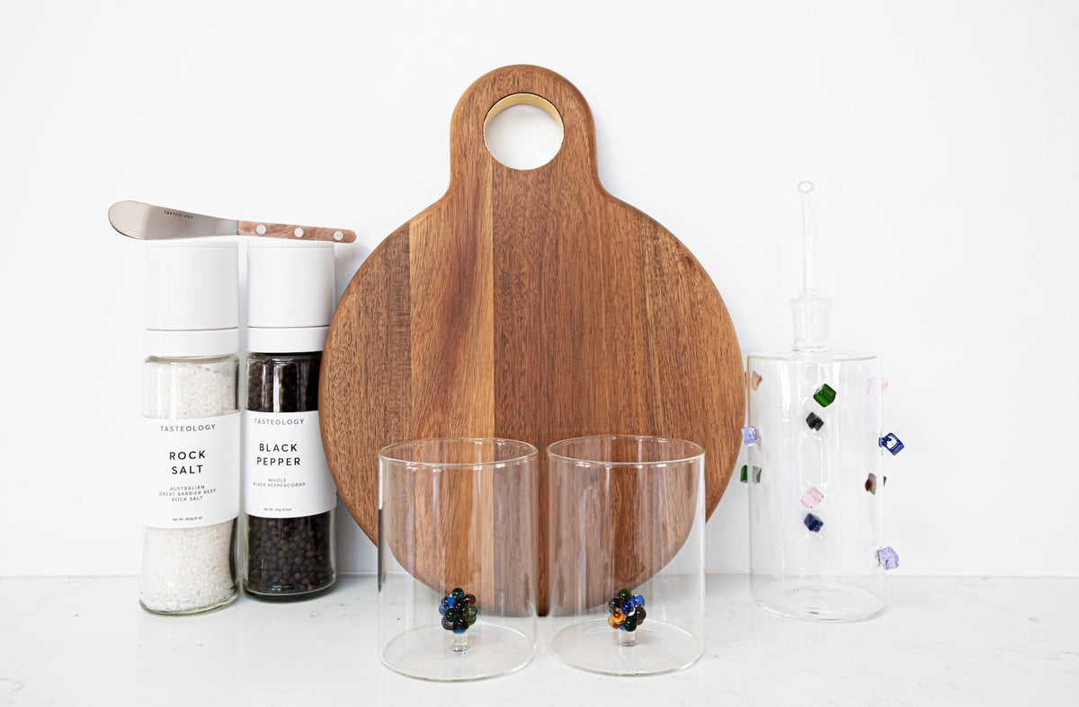 A styled kitchen or bar accessory set featuring a round wooden cutting board with a handle, two clear glass tumblers with colorful glass beads at the base, a clear glass bottle with a dropper top, and two spice grinders labeled 'ROCK SALT' and 'BLACK PEPPER'.