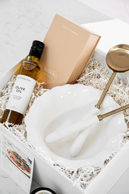 A curated gift box set featuring a bottle of olive oil, a beige branded box, a white ceramic bowl, and gold-toned serving utensils nestled in white shredded paper inside a white box.