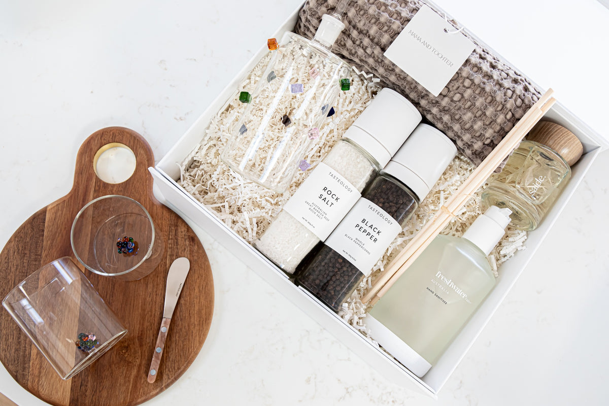 A curated gift box set from The Art of Living on a white marble surface, featuring a white box filled with spa items including black salt, white salt, a scented candle, a wooden diffuser, and a textured towel, alongside a wooden cutting board and small glass cups.