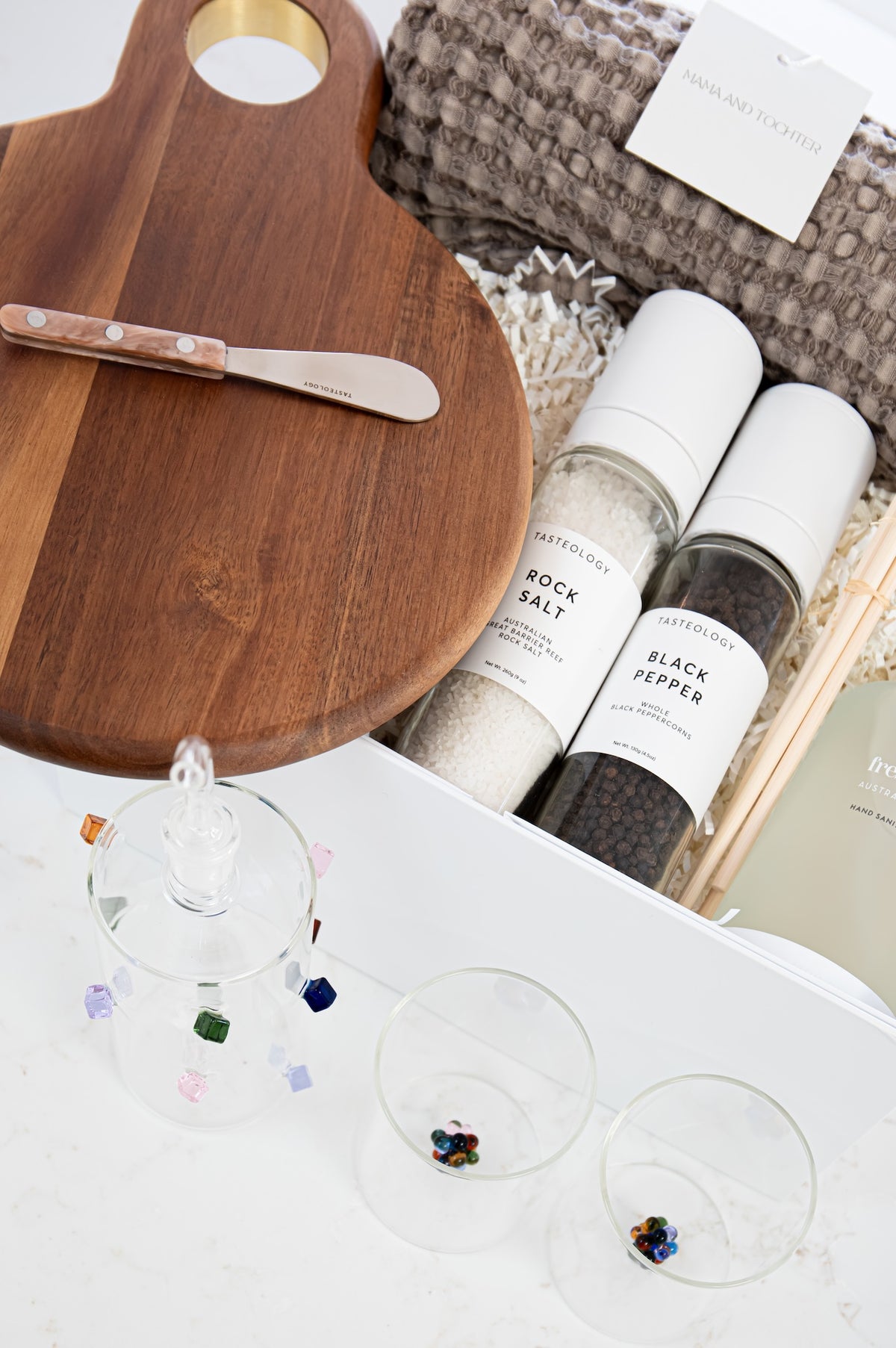 A styled gift set featuring a wooden cutting board with a small knife, two spice mills labeled 'FARMSHOP' 'SPICE SALT' and 'FARMSHOP' 'BLACK PEPPER', a knitted throw blanket, and three clear glasses with colorful gem-like accents on a white surface.