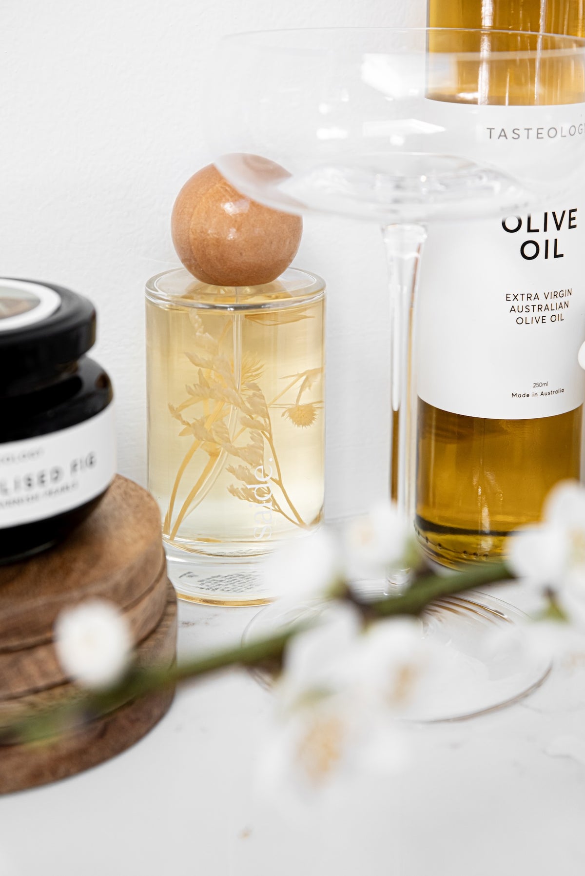 A styled product arrangement featuring a clear glass perfume bottle with a wooden stopper and etched botanical design, next to a bottle of Taste of London extra virgin Australian olive oil, with a blurred jar in the foreground on a white surface.