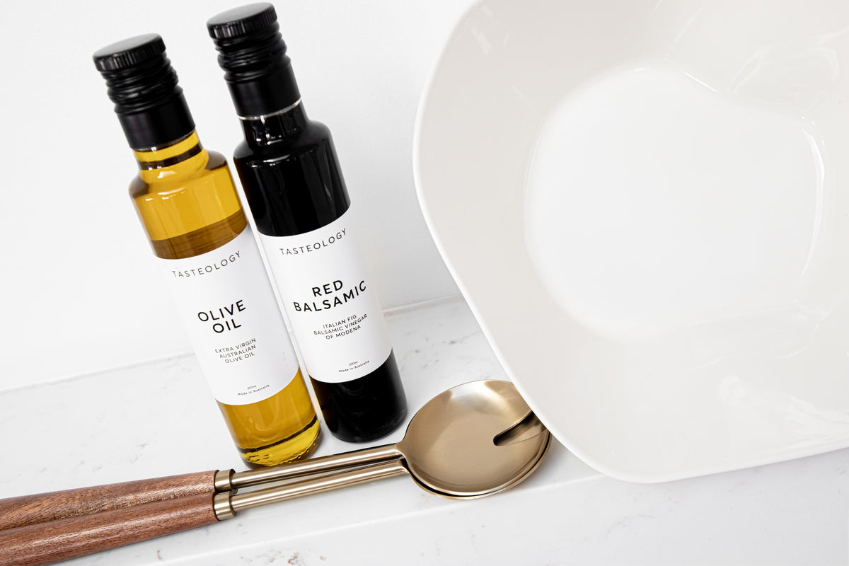 Two Tasteology bottles of olive oil and red balsamic vinegar standing on a white surface next to a large white plate and a gold spoon with a wooden handle.