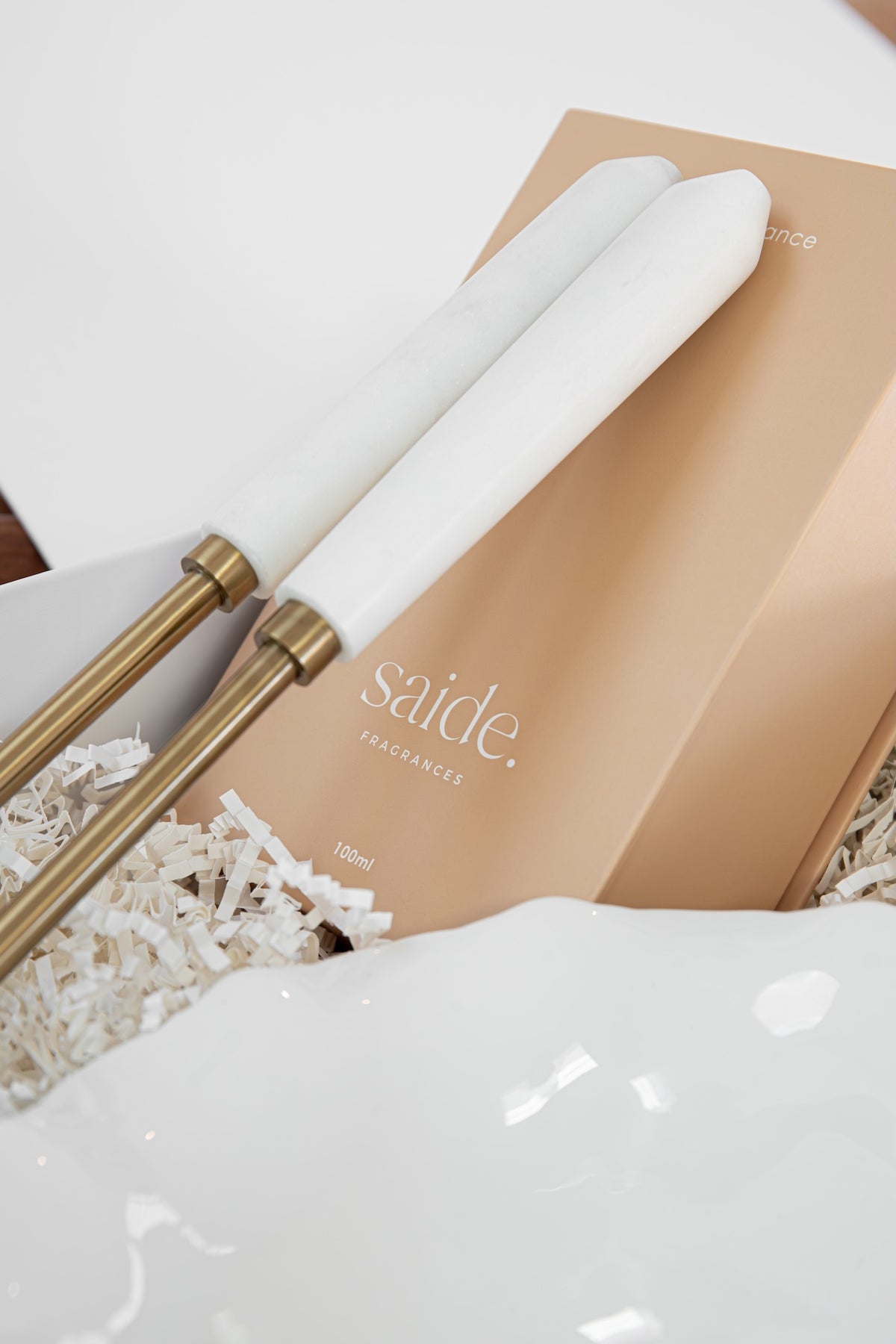 A close-up of a white marble diffuser wand with gold metal accents resting on a beige Saide Fragrances box, surrounded by white shredded paper packaging.