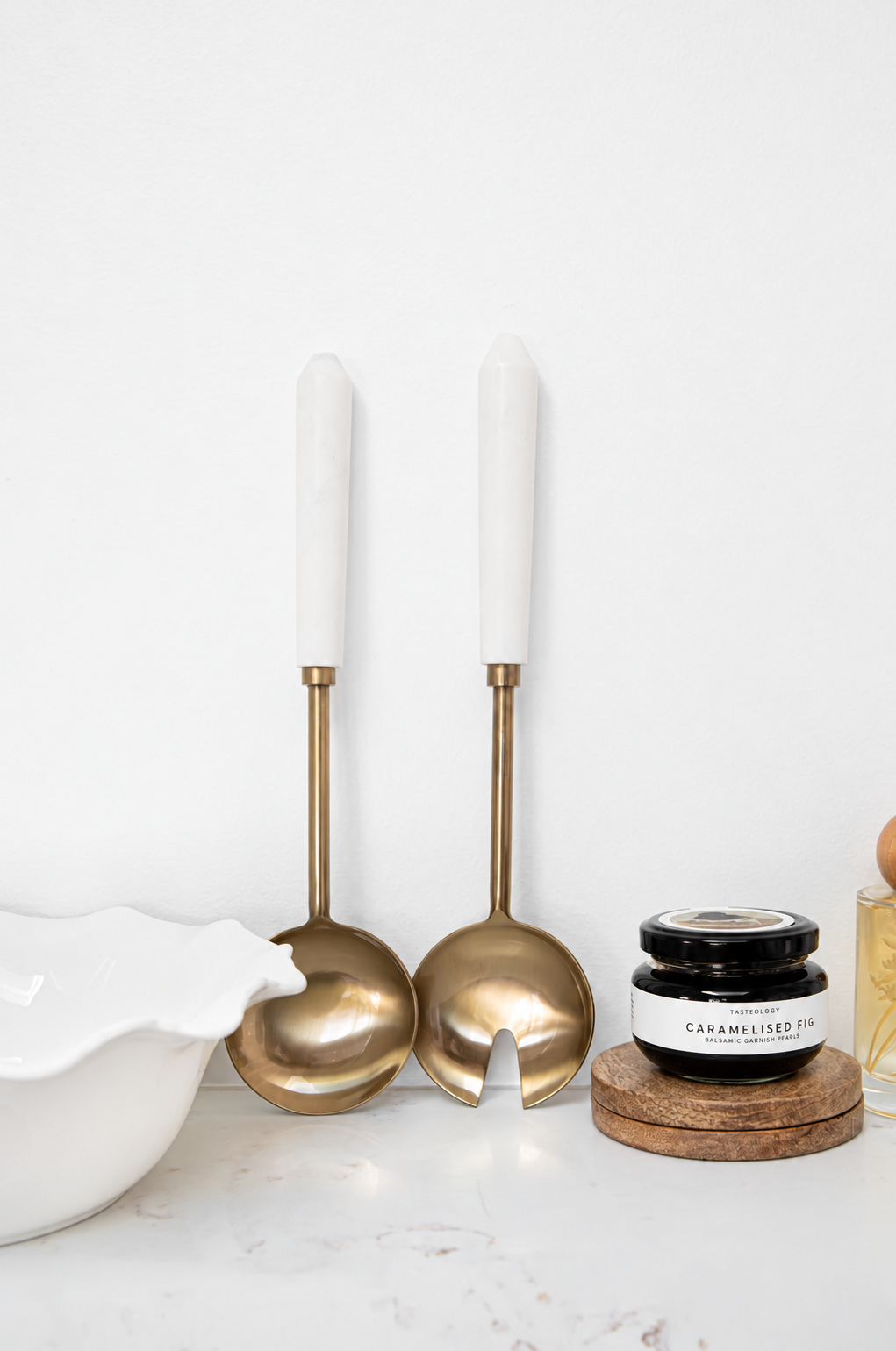 Two elegant serving utensils with white marble handles and polished gold-tone bowls, displayed on a white marble surface next to a white ceramic bowl and a small jar of caramelized tea.